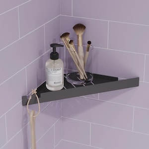 May include: A matte black triangular corner shower shelf mounted against light purple tiled walls. The shelf holds a clear pump bottle labeled L:A BRUKET and a clear glass container filled with four makeup brushes. A beige cord hangs from the front.