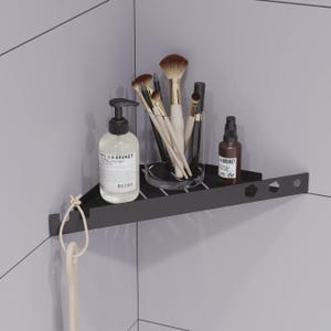 May include: A matte black triangular corner shower shelf featuring a slotted surface and geometric cutouts. It holds a clear pump bottle labeled L:A BRUKET, a glass cup filled with makeup brushes, and a small brown bottle labeled L:A BRUKET.