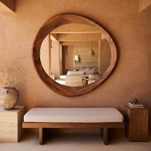 Może przedstawiać: Duże, okrągłe drewniane lustro z organiczną ramą wisi na ścianie w kolorze terakoty. Poniżej znajduje się drewniana ławka z kremową poduszką. Lustro odbija wnętrze pokoju.
