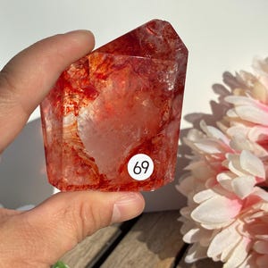 K&ouml;nnte beinhalten: Ein facettierter, rot-oranger Kristall mit inneren Adern und einem wei&szlig;en Aufkleber mit der Nummer 69. Der Kristall wird in einer Hand gehalten, mit einem verschwommenen Hintergrund aus wei&szlig;en und rosa Blumen.