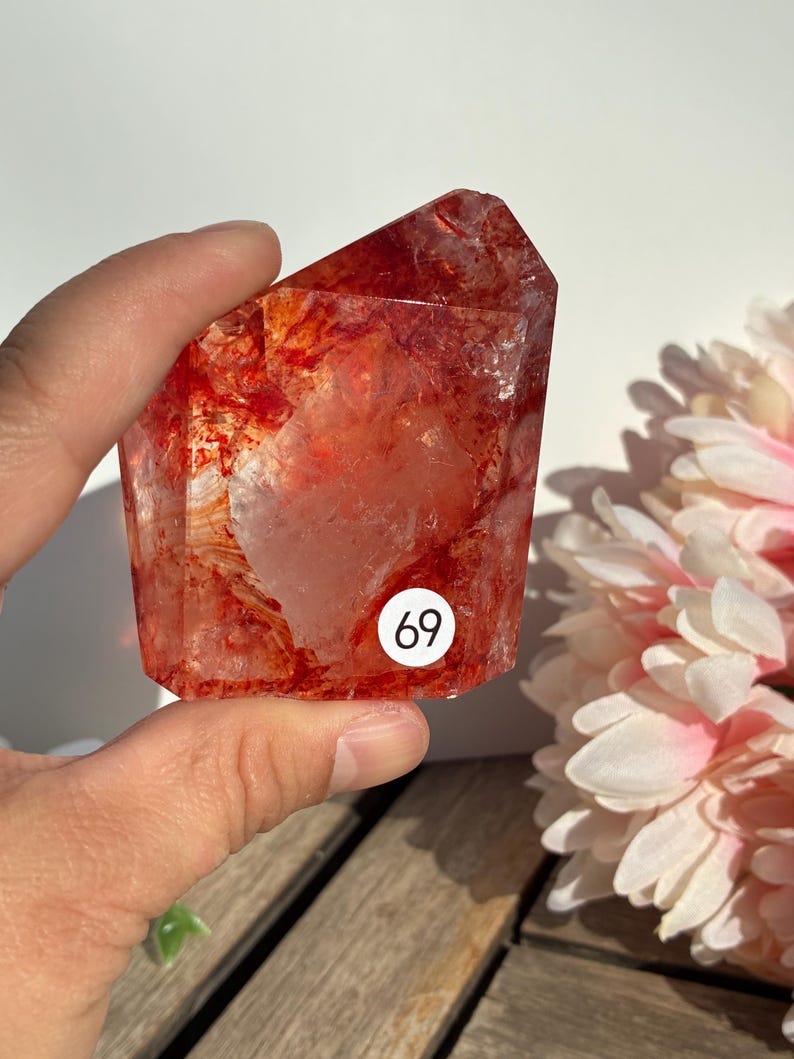 K&ouml;nnte beinhalten: Ein facettierter, rot-oranger Kristall mit inneren Adern und einem wei&szlig;en Aufkleber mit der Nummer 69. Der Kristall wird in einer Hand gehalten, mit einem verschwommenen Hintergrund aus wei&szlig;en und rosa Blumen.