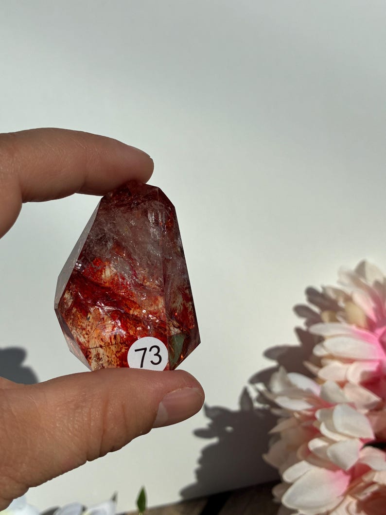 K&ouml;nnte beinhalten: Ein klarer, facettierter Kristall mit roten Einschl&uuml;ssen und einem wei&szlig;en Aufkleber mit der Nummer 73. Der Kristall wird in einer Hand vor wei&szlig;em Hintergrund gehalten, mit einer rosa-wei&szlig;en Blume im Hintergrund.