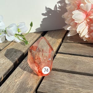 K&ouml;nnte beinhalten: Ein klarer, facettierter Kristallpunkt mit roten Einschl&uuml;ssen und einem Aufkleber mit der Nummer 74. Der Kristall liegt auf einer Holzoberfl&auml;che, mit wei&szlig;en und rosa Blumen im Hintergrund. Der Kristall hat eine r&ouml;tlich-rosa Farbe.