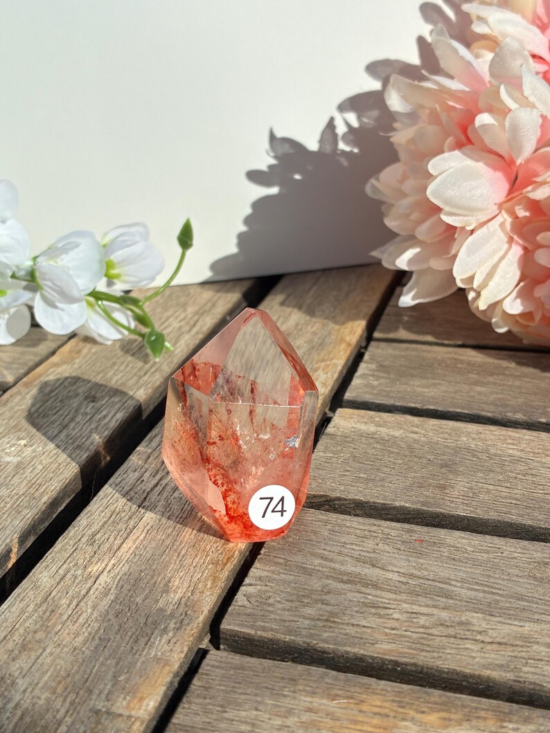 K&ouml;nnte beinhalten: Ein klarer, facettierter Kristallpunkt mit roten Einschl&uuml;ssen und einem Aufkleber mit der Nummer 74. Der Kristall liegt auf einer Holzoberfl&auml;che, mit wei&szlig;en und rosa Blumen im Hintergrund. Der Kristall hat eine r&ouml;tlich-rosa Farbe.