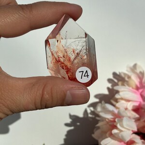 K&ouml;nnte beinhalten: Ein klarer Quarz-Kristall mit roten Einschl&uuml;ssen und einer spitzen Spitze. Der Kristall wird in einer Hand gehalten und hat einen wei&szlig;en Aufkleber mit der Nummer 74. Rosa und wei&szlig;e Blumen sind im Hintergrund.