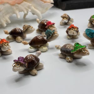 May include: A collection of small turtle figurines crafted from brown and white seashells. Each turtle features a tiny, colorful woven hat and thin gold-tone wire glasses. The turtles are arranged in a group, displaying various shell patterns and hat colors.