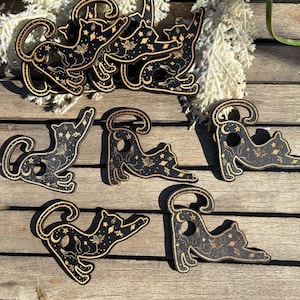 May include: A collection of black cat-shaped wooden cutouts with celestial designs. Each cat features a crescent moon, stars, and other mystical symbols. The cats are arranged on a wooden surface, with some overlapping.