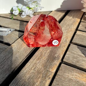 K&ouml;nnte beinhalten: Ein facettierter, rot-oranger Kristall mit inneren Einschl&uuml;ssen. Der Kristall wird auf einer verwitterten Holzoberfl&auml;che pr&auml;sentiert, mit einem kleinen wei&szlig;en Aufkleber mit der Aufschrift "70" an der Seite. Wei&szlig;e Blumen sind im Hintergrund.