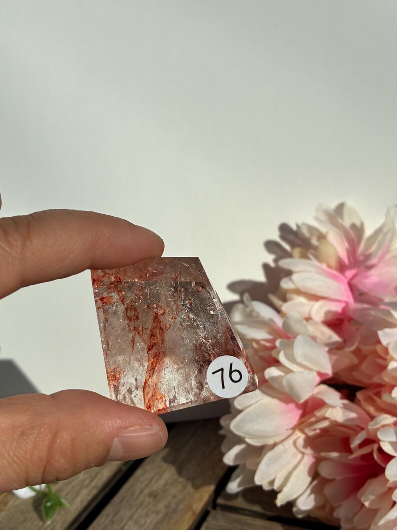K&ouml;nnte beinhalten: Ein klarer, rechteckiger Kristall mit roten Einschl&uuml;ssen und einem wei&szlig;en Aufkleber mit der Aufschrift "76". Der Kristall wird in einer Hand gehalten, mit einem verschwommenen Hintergrund aus wei&szlig;en und rosa Blumen.