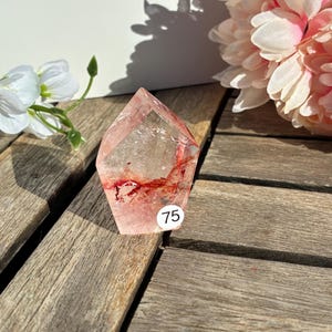 K&ouml;nnte beinhalten: Ein klarer Quarz-Kristall mit roten Einschl&uuml;ssen und einer facettierten Form. Ein wei&szlig;er Aufkleber mit der Nummer 75 befindet sich auf dem Kristall. Der Kristall liegt auf einer Holzoberfl&auml;che, mit rosa und wei&szlig;en Blumen im Hintergrund.