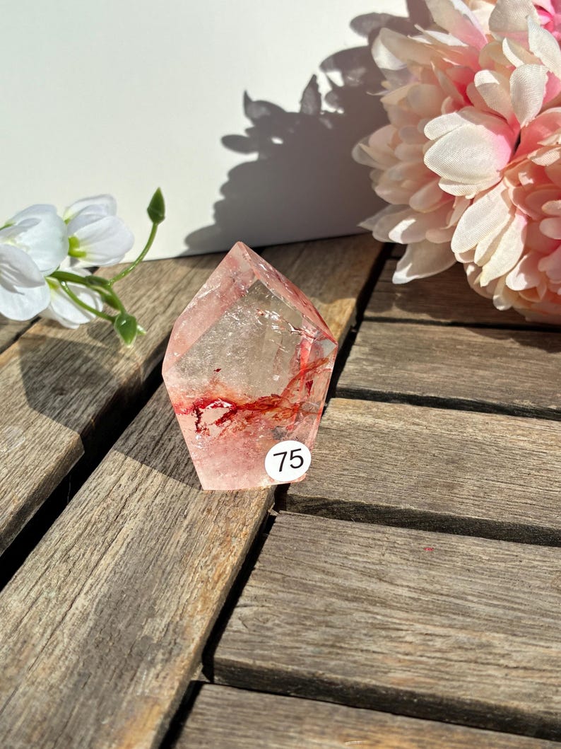 K&ouml;nnte beinhalten: Ein klarer Quarz-Kristall mit roten Einschl&uuml;ssen und einer facettierten Form. Ein wei&szlig;er Aufkleber mit der Nummer 75 befindet sich auf dem Kristall. Der Kristall liegt auf einer Holzoberfl&auml;che, mit rosa und wei&szlig;en Blumen im Hintergrund.