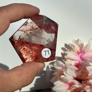 K&ouml;nnte beinhalten: Ein klarer, facettierter Kristall mit roten Einschl&uuml;ssen und einem Aufkleber mit der Aufschrift "71". Der Kristall wird in einer Hand gehalten, mit rosa und wei&szlig;en Blumen im Hintergrund. Die Form des Kristalls ist geometrisch.