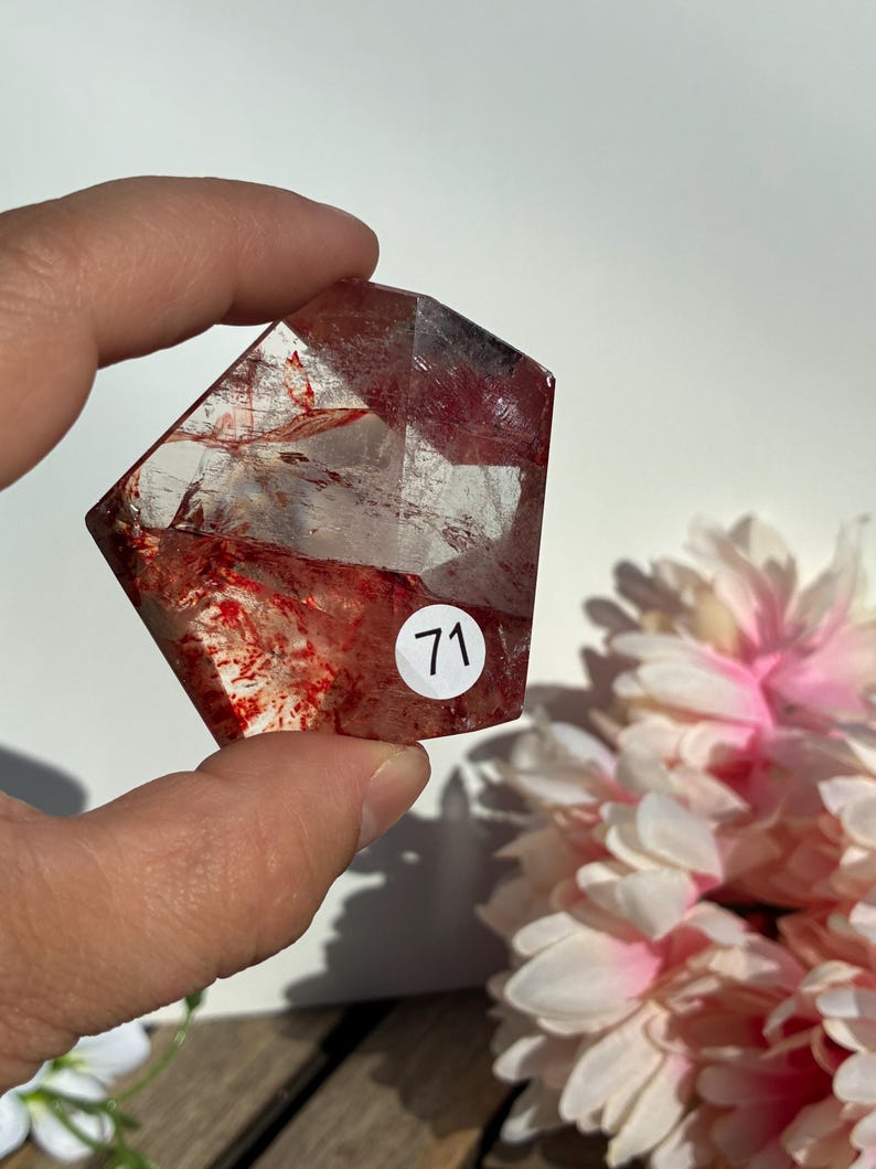 K&ouml;nnte beinhalten: Ein klarer, facettierter Kristall mit roten Einschl&uuml;ssen und einem Aufkleber mit der Aufschrift "71". Der Kristall wird in einer Hand gehalten, mit rosa und wei&szlig;en Blumen im Hintergrund. Die Form des Kristalls ist geometrisch.