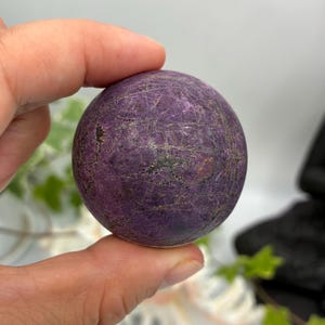 May include: A spherical stone with a deep purple hue, featuring a complex network of dark, intersecting veins across its textured surface. The orb displays natural variations in color and pattern, with a matte finish and a solid, rounded form.