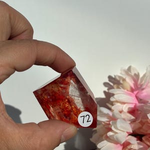 K&ouml;nnte beinhalten: Ein facettierter, rot-oranger Kristall mit inneren Einschl&uuml;ssen wird in einer Hand gehalten. Ein wei&szlig;er Aufkleber mit der Nummer 72 befindet sich auf dem Kristall. Rosa und wei&szlig;e Blumen sind im Hintergrund. Der Kristall ist ein Edelstein.