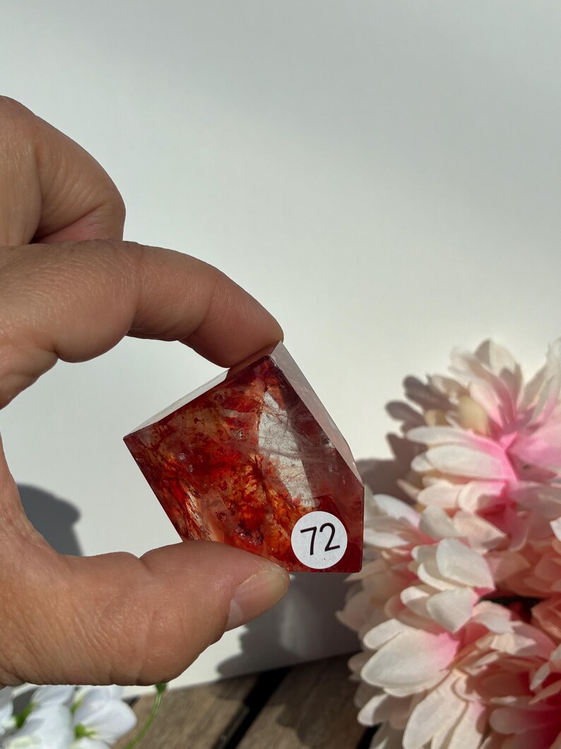 K&ouml;nnte beinhalten: Ein facettierter, rot-oranger Kristall mit inneren Einschl&uuml;ssen wird in einer Hand gehalten. Ein wei&szlig;er Aufkleber mit der Nummer 72 befindet sich auf dem Kristall. Rosa und wei&szlig;e Blumen sind im Hintergrund. Der Kristall ist ein Edelstein.