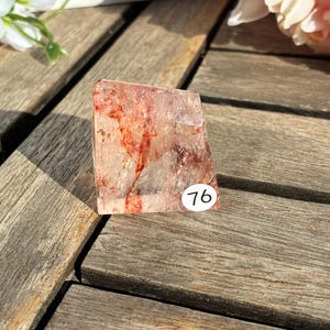 K&ouml;nnte beinhalten: Ein klarer Quarz-Kristall mit roten Einschl&uuml;ssen und einem kleinen wei&szlig;en Aufkleber mit der Nummer 76. Der Kristall ist rechteckig und steht auf einer Holzoberfl&auml;che. Der Kristall wird von Sonnenlicht beleuchtet.