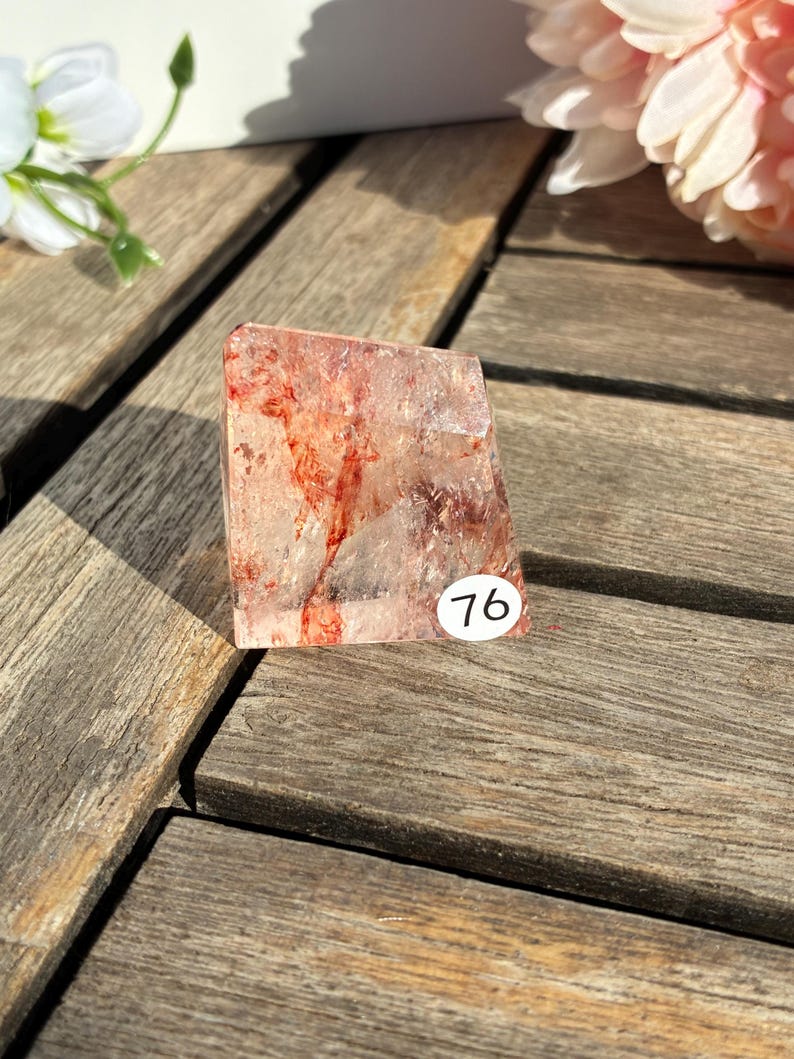 K&ouml;nnte beinhalten: Ein klarer Quarz-Kristall mit roten Einschl&uuml;ssen und einem kleinen wei&szlig;en Aufkleber mit der Nummer 76. Der Kristall ist rechteckig und steht auf einer Holzoberfl&auml;che. Der Kristall wird von Sonnenlicht beleuchtet.