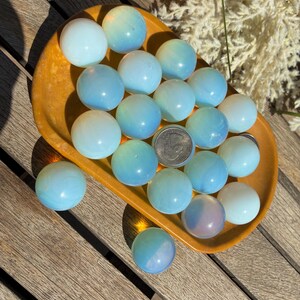 May include: A collection of opalescent spheres in shades of blue and white, displayed in a small, orange-brown dish. The spheres vary in size and color, with some showing hints of iridescence. The arrangement is on a wooden surface, with a coin visible.
