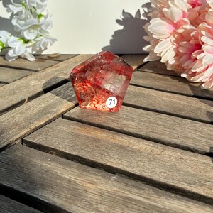 K&ouml;nnte beinhalten: Ein facettierter, rotbrauner Kristall mit inneren Einschl&uuml;ssen, der auf einer Holzoberfl&auml;che liegt. Der Kristall hat einen kleinen wei&szlig;en Aufkleber mit der Nummer 71. Wei&szlig;e und rosa Blumen sind im Hintergrund.