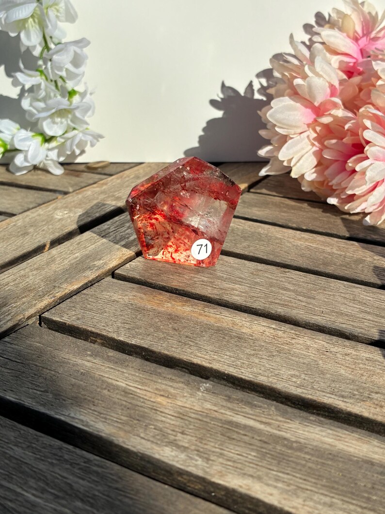 K&ouml;nnte beinhalten: Ein facettierter, rotbrauner Kristall mit inneren Einschl&uuml;ssen, der auf einer Holzoberfl&auml;che liegt. Der Kristall hat einen kleinen wei&szlig;en Aufkleber mit der Nummer 71. Wei&szlig;e und rosa Blumen sind im Hintergrund.