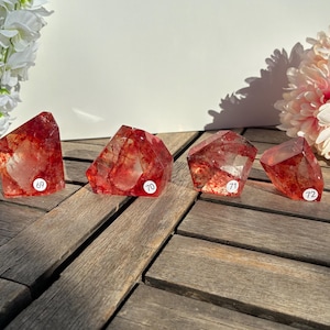 K&ouml;nnte beinhalten: Vier facettierte rote Quarz-Kristalle mit inneren Einschl&uuml;ssen, jeweils mit einem wei&szlig;en Aufkleber mit der Nummer 69, 70, 71 und 72. Die Kristalle werden auf einer verwitterten Holzoberfl&auml;che mit Blumenarrangements im Hintergrund pr&auml;sentiert.