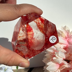 K&ouml;nnte beinhalten: Ein klarer Quarz-Kristall mit roten Einschl&uuml;ssen, gehalten in einer Hand. Der Kristall ist facettiert mit einer geometrischen Form. Ein wei&szlig;er Aufkleber mit der Nummer 70 ist sichtbar. Rosa und wei&szlig;e Blumen sind im Hintergrund.