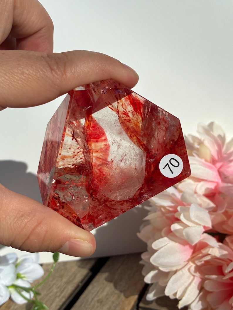 K&ouml;nnte beinhalten: Ein klarer Quarz-Kristall mit roten Einschl&uuml;ssen, gehalten in einer Hand. Der Kristall ist facettiert mit einer geometrischen Form. Ein wei&szlig;er Aufkleber mit der Nummer 70 ist sichtbar. Rosa und wei&szlig;e Blumen sind im Hintergrund.