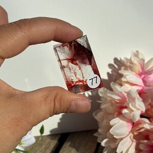 K&ouml;nnte beinhalten: Rechteckiger, klarer Harzanh&auml;nger mit roten und wei&szlig;en Einschl&uuml;ssen. Der Anh&auml;nger hat die Nummer "77" auf einem wei&szlig;en Aufkleber. Der Hintergrund umfasst rosa und wei&szlig;e Blumen und eine Holzoberfl&auml;che.
