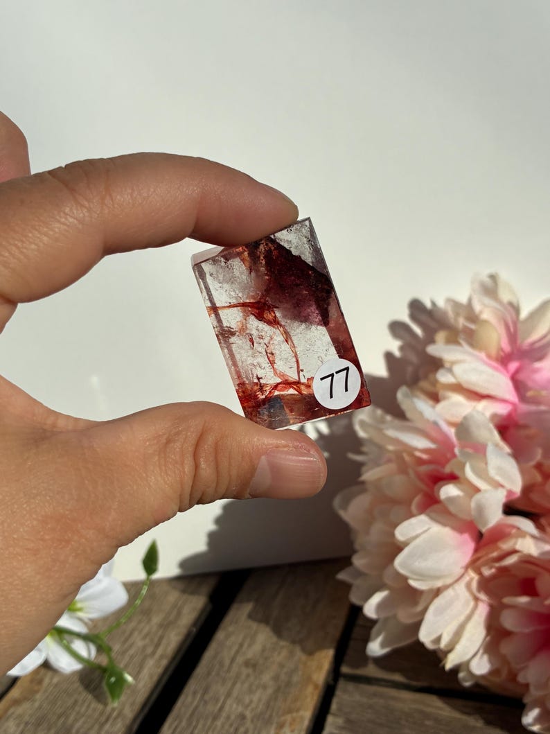 K&ouml;nnte beinhalten: Rechteckiger, klarer Harzanh&auml;nger mit roten und wei&szlig;en Einschl&uuml;ssen. Der Anh&auml;nger hat die Nummer "77" auf einem wei&szlig;en Aufkleber. Der Hintergrund umfasst rosa und wei&szlig;e Blumen und eine Holzoberfl&auml;che.