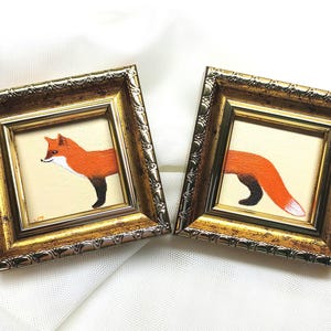 May include: Two small framed paintings. One shows a fox from the side, with orange fur, black legs, and a white chest. The other shows the fox's tail. The frames are gold with ornate detailing.