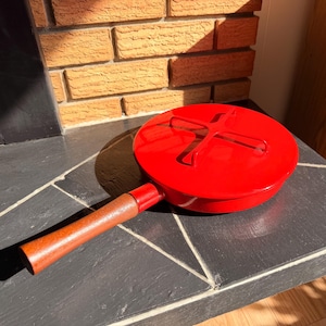 Puede incluir: Una sartén roja brillante con un mango de madera. La sartén es redonda con una tapa que tiene un diseño en forma de cruz. El mango es de color marrón oscuro. La sartén está sobre una superficie gris oscura.