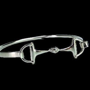 May include: A silver-colored bangle bracelet featuring a horse bit design. The bracelet has a curved, slender band with the bit centered, showcasing a classic equestrian theme. The metal has a polished, reflective finish.