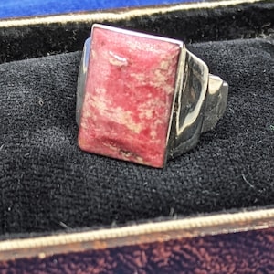 May include: A silver ring featuring a rectangular, flat-topped stone with a mottled pink and white surface. The metal band is polished and smooth, holding the stone securely in a bezel setting. The ring is displayed in a dark, textured case.