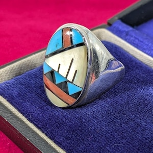 May include: A silver ring featuring an oval face with inlaid geometric segments of turquoise, coral, and mother-of-pearl. The inlay is separated by thin black lines, creating a mosaic pattern on the polished metal band with a smooth finish.