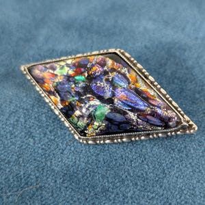 Op de afbeelding: Een ruitvormige broche met een donkere, iriserende glazen inleg met levendige spikkels in blauw, paars, oranje en groen. Het glas is gevat in een getextureerd, zilverkleurig metalen frame met een kralenrand langs de omtrek.