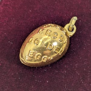 May include: A gold-coloured metal charm in the shape of an egg with a small loop at the top. The surface of the egg features the embossed text MECCA GOLDEN EGG arranged in three lines across the centre of the oval-shaped object.