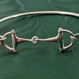 May include: A polished silver-tone bangle bracelet featuring a central horse bit design. The metal band is smooth and circular, connecting to the equestrian-inspired bit hardware that serves as the focal point of this jewelry piece.