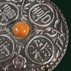 May include: Close-up of a silver-toned, circular decorative object with an amber-coloured, round stone at the centre. The object features four Chinese characters in circular frames, surrounded by floral and bamboo-like patterns.