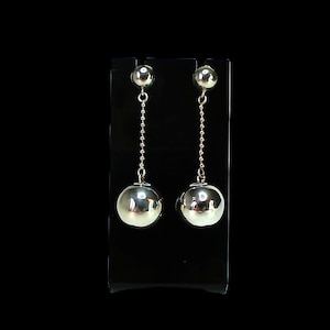 May include: A pair of silver-colored drop earrings. Each earring features a small ball stud, a delicate chain, and a larger, polished sphere. The earrings are displayed against a black background, highlighting their reflective surfaces.