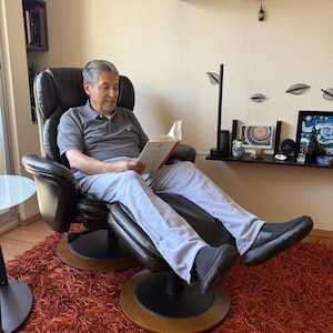 Op de afbeelding: Een donkerbruine leren relaxfauteuil met een bijpassende ronde voetenbank. De stoel heeft een hoge rugleuning, gevoerde armleuningen en een draaibare voet met een houten afwerking. De fauteuil staat op een getextureerd oranje tapijt.