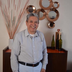 May include: A person wearing a light gray button-down shirt with a subtle vertical stripe pattern, a black leather belt with a gold buckle, and light blue trousers. The person is smiling while standing in front of a dark wood cabinet.