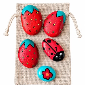 May include: A beige burlap drawstring pouch containing five painted stones. Two stones are red with yellow dots and teal tops, one is red with yellow dots and a teal top, one is red with black spots and a black head, and one is blue with a red flower.