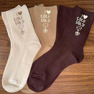 May include: Three pairs of ribbed crew socks in cream, tan, and burgundy. Each sock features the text "I ❤️ Cocktails" with a cocktail glass graphic. The socks are displayed on a wooden surface.