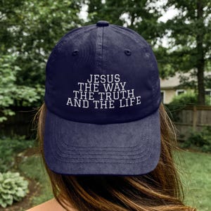 May include: A navy blue baseball cap featuring white embroidered text on the front that reads JESUS THE WAY THE TRUTH AND THE LIFE. The cap has a structured crown, a curved brim with visible stitching, and a button at the top centre of the crown.