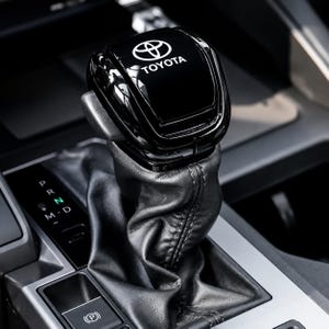 May include: A black gear shift knob featuring the white Toyota logo on top. The knob is attached to a black leather boot with visible stitching. To the left, a gear indicator panel displays the letters P, R, N, D, M, and a plus and minus sign.