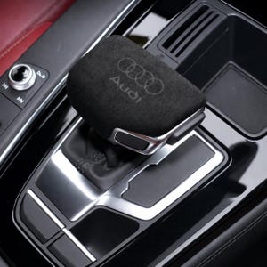 May include: A black suede gear shift knob cover featuring the Audi logo embossed on the top surface. The cover is fitted onto a vehicle gear shifter surrounded by a black console with silver metallic trim accents and a circular control dial.