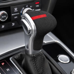 Könnte beinhalten: Ein Auto-Schaltknauf mit einer schwarzen, strukturierten, wildlederartigen Oberseite mit einem horizontalen roten Streifen, einer silbernen Metallseitenblende und einer schwarzen Lederbasis mit sichtbaren Nähten in der Mittelkonsole.