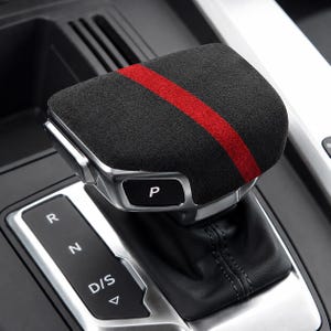 May include: A black suede gear shift knob cover featuring a prominent red stripe across the top. The cover is installed on a vehicle gear shifter with a silver metallic side panel, a black P button, and a black leather boot with visible stitching.