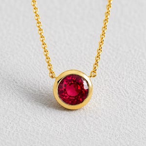May include: A gold chain necklace featuring a single round, faceted red gemstone set in a smooth, polished gold bezel setting. The vibrant red stone is centred on the delicate gold link chain, creating a simple and elegant piece of jewellery.